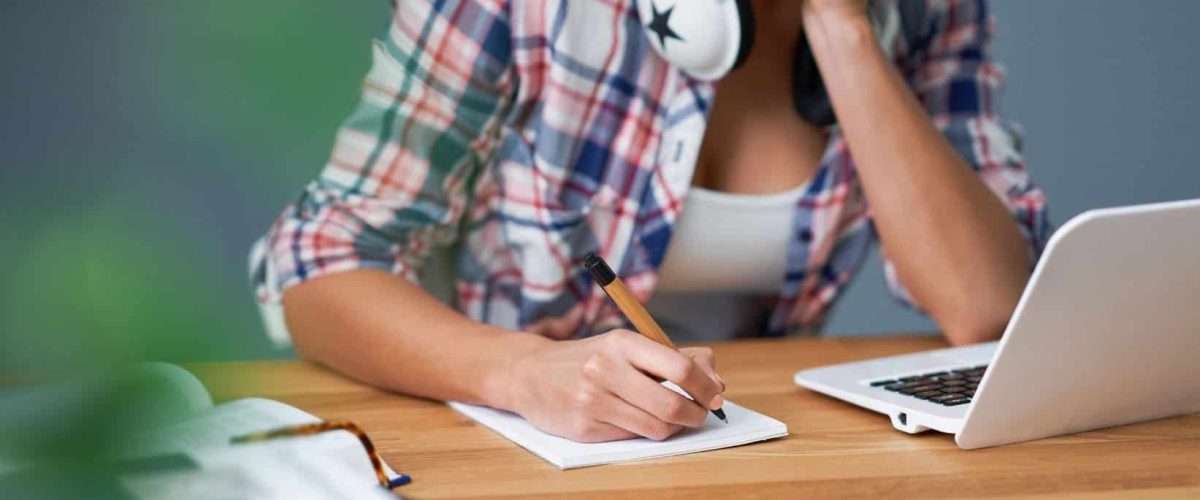 Self Paced Online Learning- Female In Shirt Wearing Headphones Around Her Neck Sitting In Front Of A Laptop And A Notepad And Pen